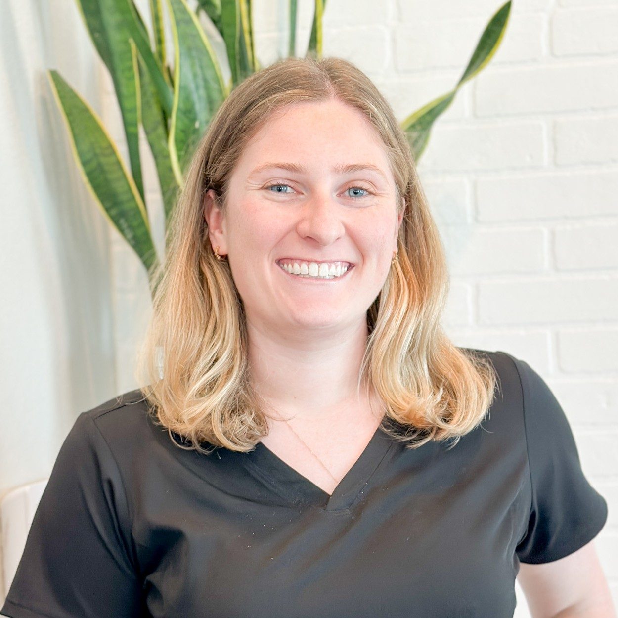Chiropractic Assistant Katy TX Katie Meet The Team Katie standing in front of white brick wall and plant smiling and wearing a black top.